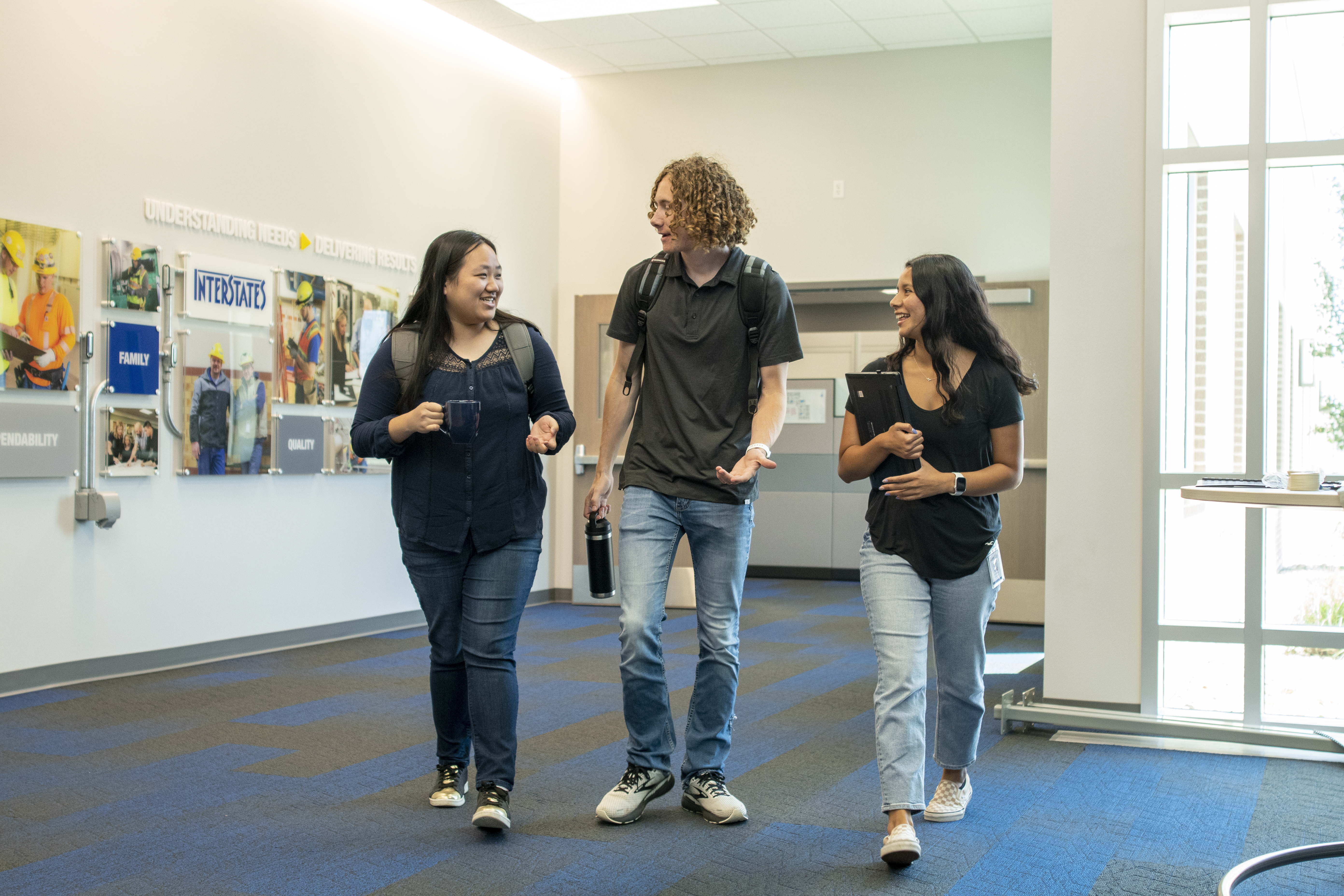 Three students walking and chatting in office hallway with Interstate company branding and photos displayed on wall