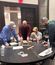 Several people work together on a tabletop building challenge near a doorway in a conference room.