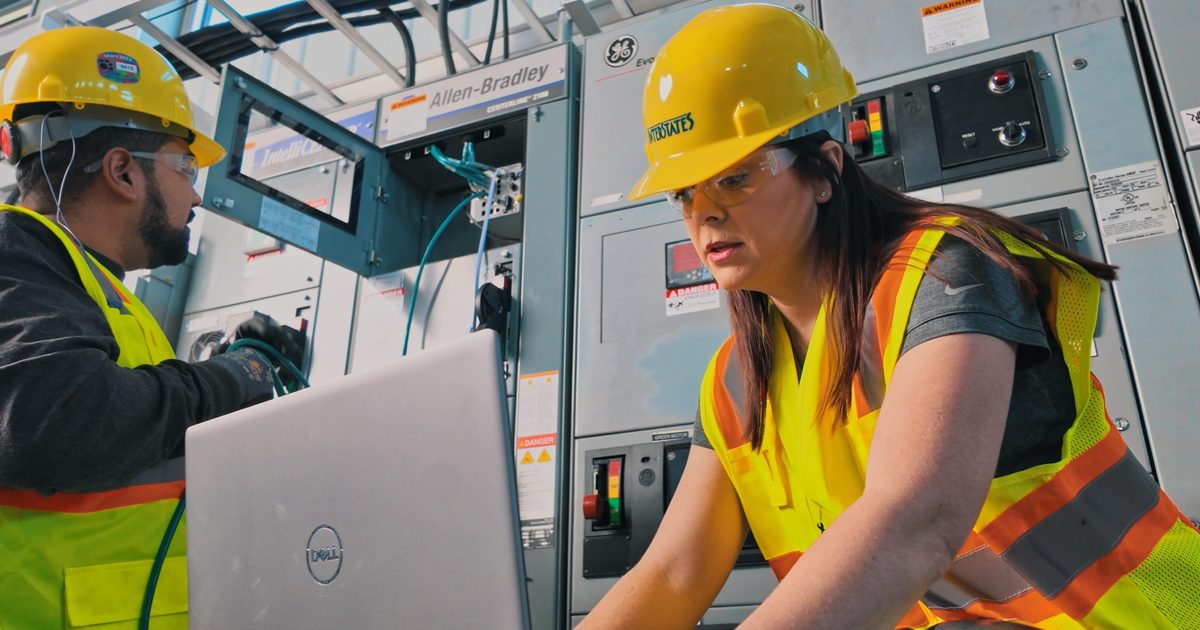 Individual on a plant floor using her computer while a man works with network wires in an industrial room.