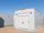 A prefabricated electrical enclosure (E-House) installed outdoors on a prepared site under a clear blue sky. The white modular building features a secure entry door, exterior lighting, and integrated HVAC equipment for climate control.
