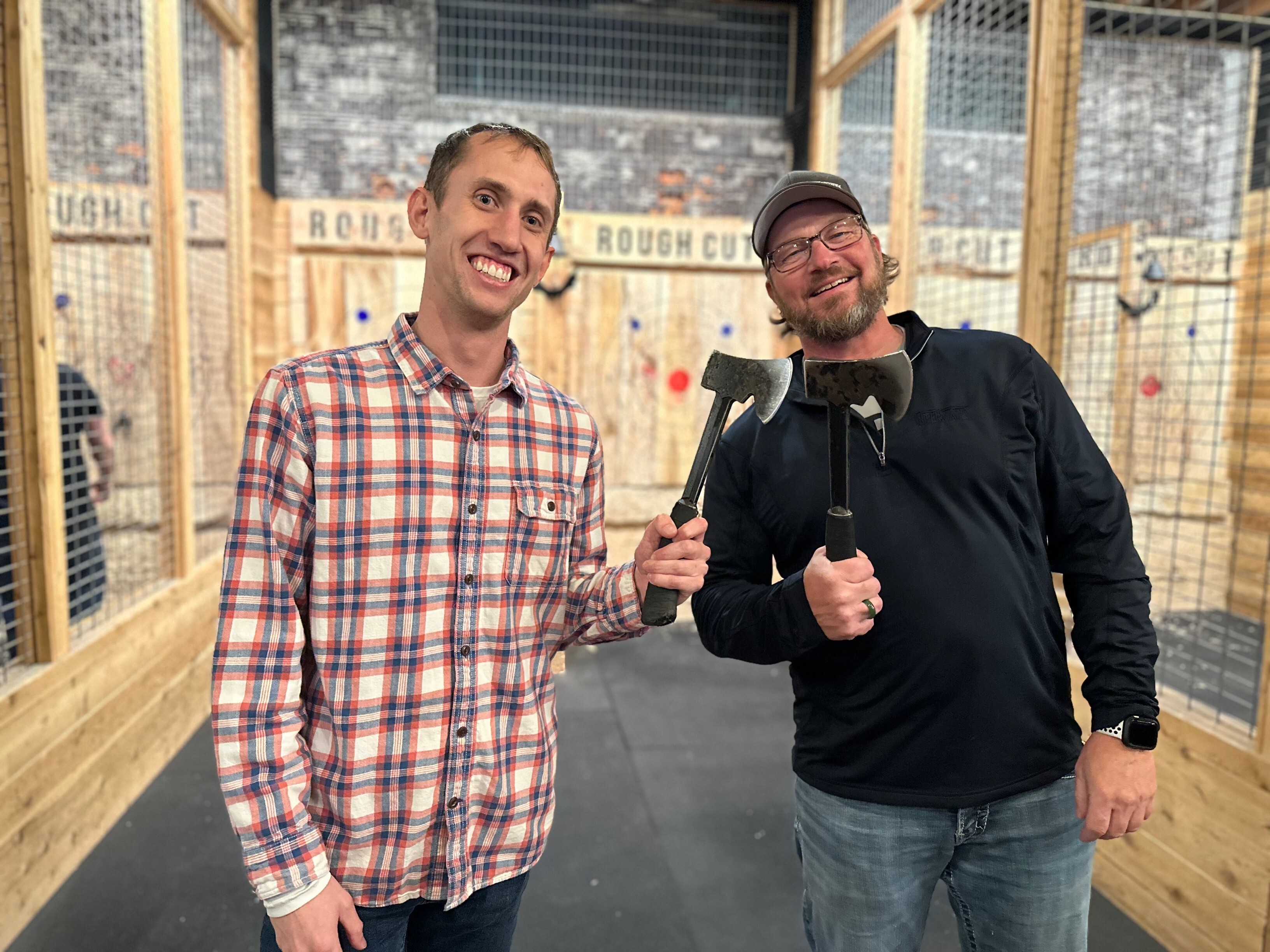 Two men smiling and holding axes at an axe-throwing range, with wooden targets visible in the background.