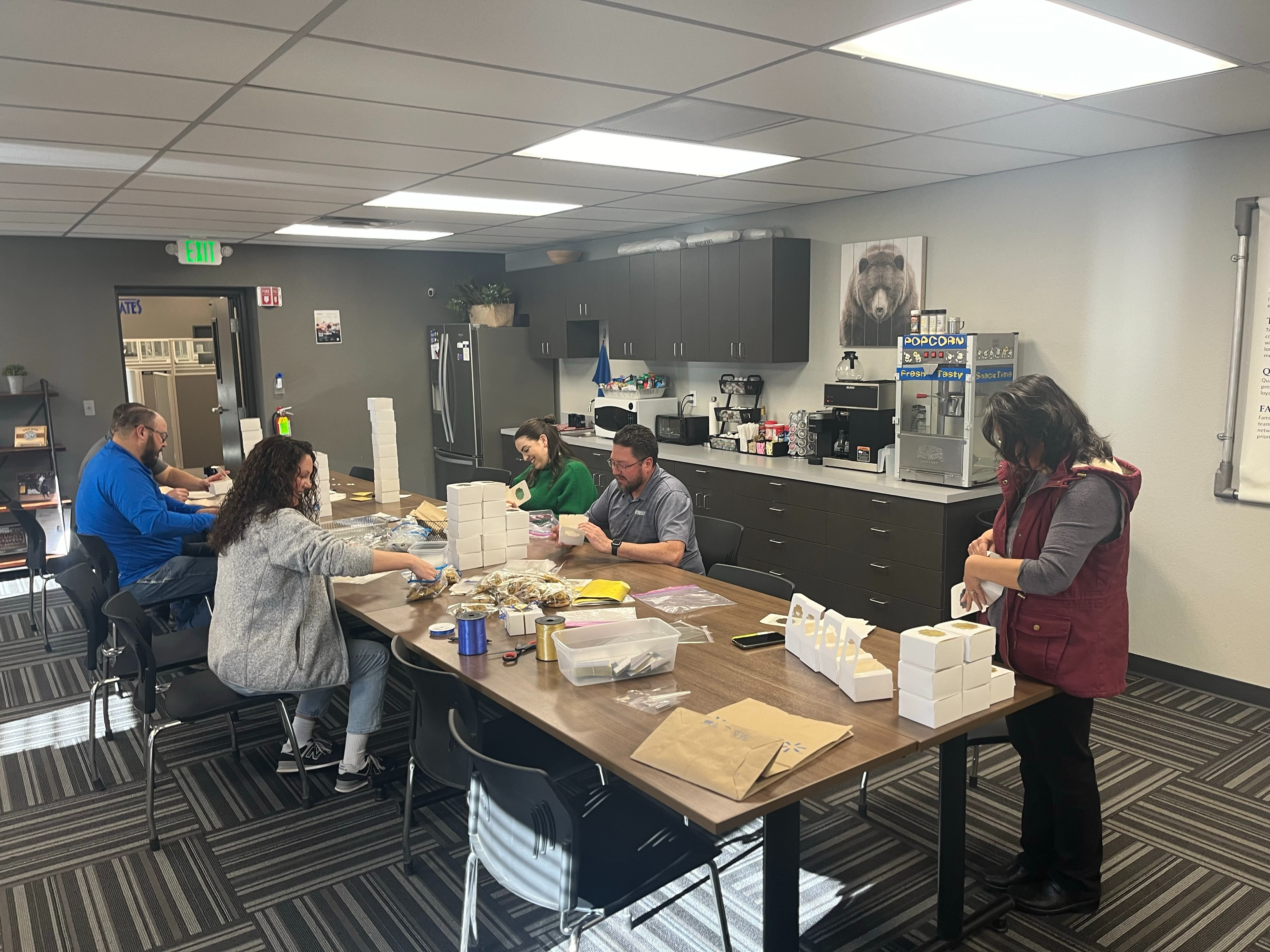 Office break room scene showing employees around table assembling or packaging items together, with kitchen amenities and popcorn machine visible in background
