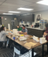 Office break room scene showing employees around table assembling or packaging items together, with kitchen amenities and popcorn machine visible in background