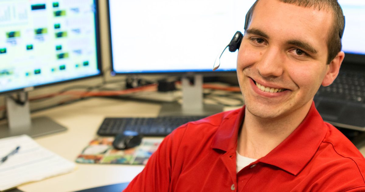 Man in a red shirt in front of monitors with his arms crossed. He has on a headset to help with support calls.