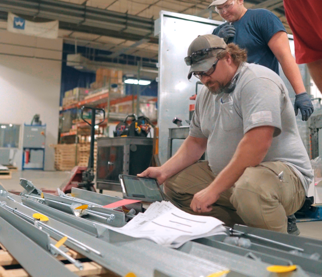 Three workers in a fabrication shop kneel and stand over a metal assembly laid out on a pallet, reviewing printed plans and a digital tablet to guide installation.