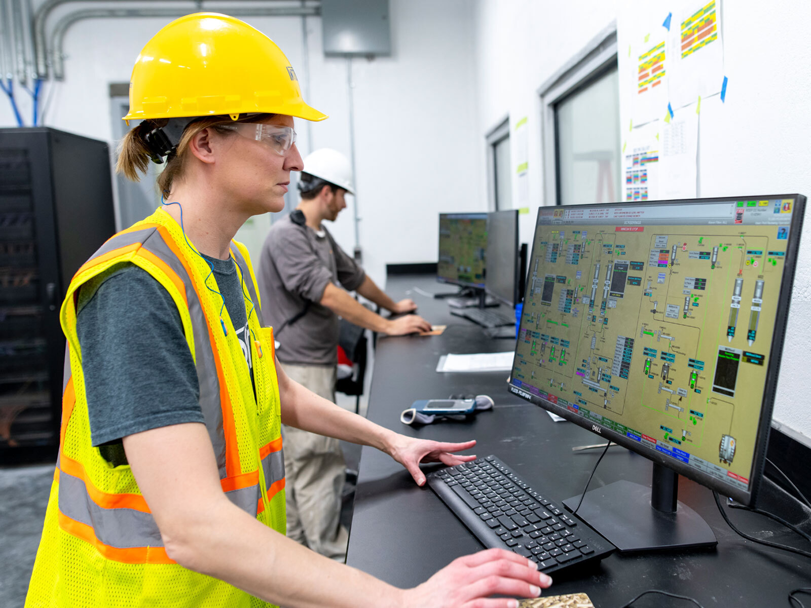 A woman wearing a yellow safety vest and hard hat operates a control system at a computer station, while another worker focuses on a separate monitor in the background.
