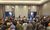 A large group of people wearing casual clothes and work hats gathers in a hotel conference room, facing a small setup at the front where a few people appear to be speaking or giving instructions.