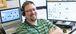 Man wearing a green shirt smiling in front of two computer monitors.