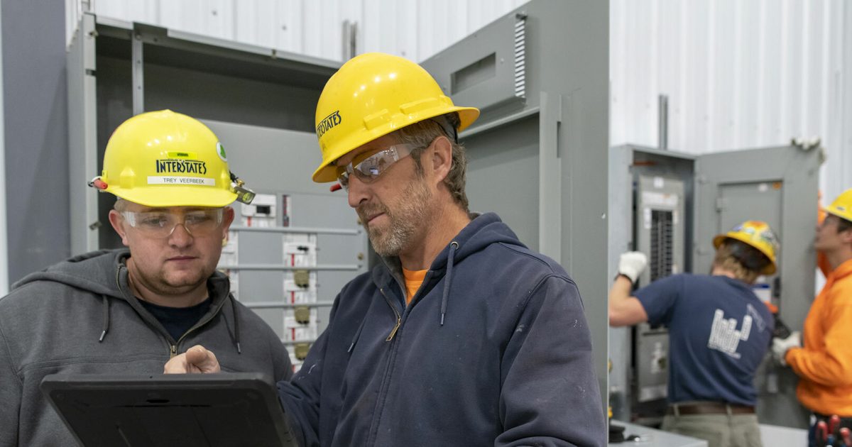 Two men wearing yellow hardhats viewing something on a tablet.