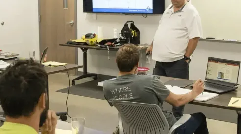 An instructor leads a small classroom of apprentices, presenting electrical concepts on a screen and whiteboard while participants follow along with notes and laptops.