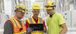 Three men wearing PPE gear viewing something on an iPad.