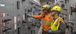 Two men wearing PPE in an electric room.