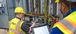 A photo of two workers wearing safety gear, including yellow hard hats, high-visibility vests with "Interstates" logos, safety glasses, and gloves, working on a large control panel filled with wiring and electronic components.