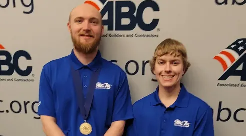 Two individuals wearing blue ABC polo shirts stand in front of an ABC backdrop, smiling at the camera, with one person wearing a medal around their neck.