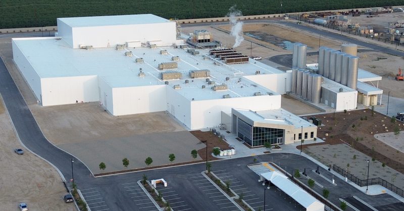 Aerial view of a large, modern industrial facility with white roofing, multiple connected buildings, and several tall stainless steel storage tanks. The site includes landscaped areas, a main entrance with a glass façade, a large parking lot, and surrounding open land.
