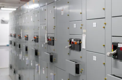 Interior of an electrical e-house showing a long row of gray motor control center (MCC) cabinets with labeled panels, switches, and indicator lights, in a clean, well-lit industrial space.