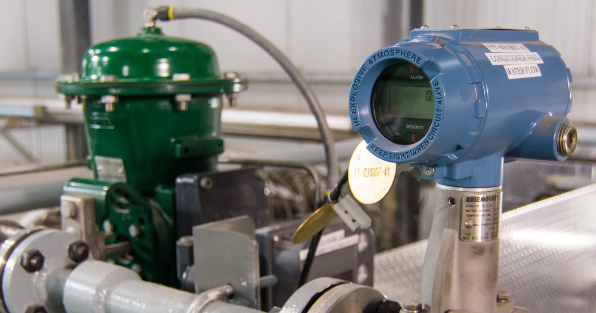 Close-up of industrial piping inside a manufacturing facility, featuring a blue digital flow transmitter mounted on a metal pipe in the foreground and a green control valve actuator behind it.