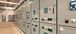 A row of industrial electrical control panels lines the wall of a clean, well-lit facility, with labeled switches, digital displays, and control knobs visible on each unit.