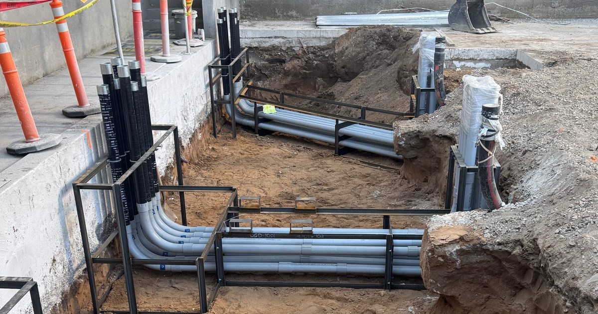 A construction site shows an underground electrical installation in progress. Several gray PVC conduits are neatly arranged in rows within a rectangular trench, supported by black metal framing to keep them evenly spaced.