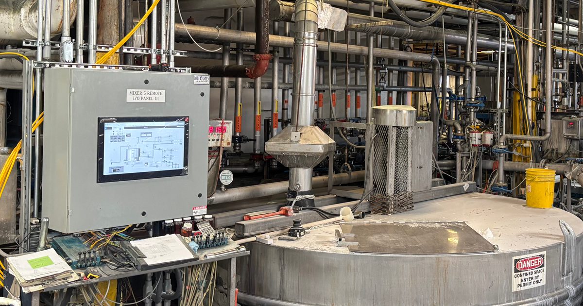 Industrial processing area with a large stainless steel mixing tank surrounded by extensive piping, valves, and conduit.