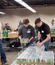 Workers stack shrink-wrapped cases of canned drinks on a pallet in a warehouse, with a forklift and other supplies visible in the background.