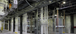 A clean and modern industrial electrical room with polished concrete floors and multiple vertical bus duct systems enclosed in metal housings. Overhead, a dense network of metal electrical conduit and cable trays is neatly organized, supplying power and control to the machinery below.
