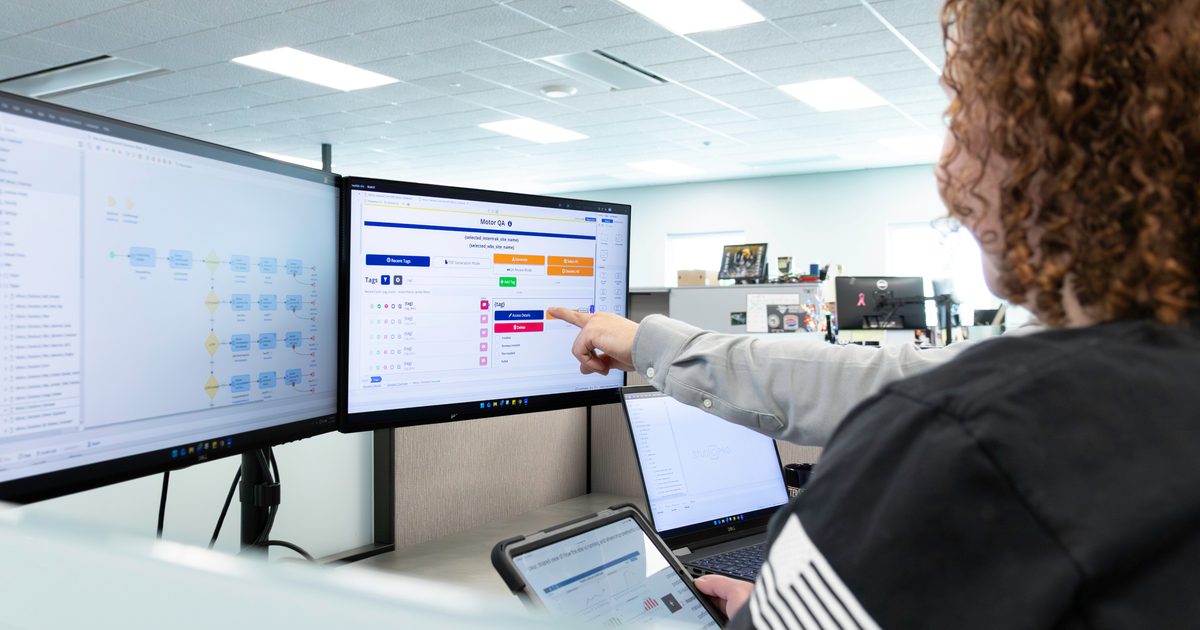 A person in an office points at a computer monitor displaying a workflow interface while working across multiple screens.