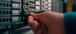 A close-up of a person’s hand installing a component in a server rack, with multiple hard drive bays and network equipment visible.