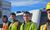 Four construction workers stand side by side in front of a white truck. They are wearing safety vests and gear. Behind them is a mobile office.