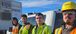 Four construction workers stand side by side in front of a white truck. They are wearing safety vests and gear. Behind them is a mobile office.
