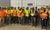 A group of construction workers in safety vests and hard hats stands inside a large industrial building. Several workers are wearing bright orange and yellow high-visibility gear, and one person in the center is holding a safety award plaque.
