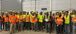 Large group of construction workers wearing high-visibility vests, yellow hard hats, and safety glasses pose indoors; one holds a championship-style belt.