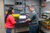 Two people stand in a technical training lab or workspace, collaborating at a workbench with laptops. One person gestures toward a computer screen while explaining something, and the other listens while holding a keyboard.