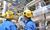 Two industrial workers in blue uniforms and yellow hard hats, equipped with hearing protection, stand in front of complex metal piping and large equipment at a processing or manufacturing plant. One worker is pointing towards a large stainless steel vessel while the other observes. The background shows multiple pipes, tanks, and safety railings, indicating a highly engineered industrial environment.