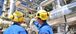 Two industrial workers in blue uniforms and yellow hard hats, equipped with hearing protection, stand in front of complex metal piping and large equipment at a processing or manufacturing plant. One worker is pointing towards a large stainless steel vessel while the other observes. The background shows multiple pipes, tanks, and safety railings, indicating a highly engineered industrial environment.