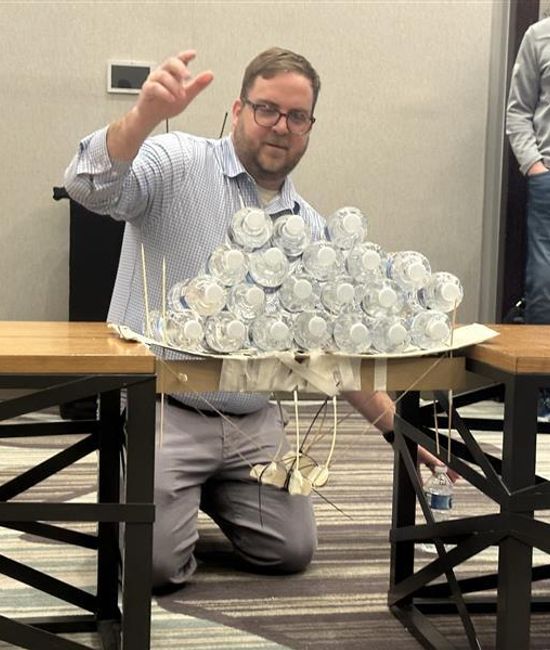 Man kneeling beside a tabletop structure made from stacked water bottles during a team activity.