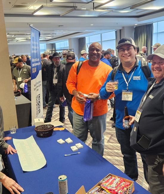 Three men and two women stand at a booth in a conference room, smiling and talking with attendees.