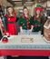 Three women in festive Christmas costumes stand behind a holiday-themed table at an office party, with a decorated gingerbread house in the foreground.