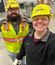 Two workers in yellow hard hats and safety vests smile for a selfie inside a large industrial workspace.
