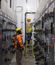 Two electricians wearing safety gear work inside a narrow industrial electrical room lined with large gray control panels on both sides. One worker stands on a ladder adjusting equipment on the right panel, while the other observes from the floor.