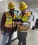 Two construction workers in hard hats and safety vests stand in a hallway, reviewing information on a tablet together.