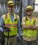 Two construction workers wearing safety vests, hard hats, gloves, and safety glasses stand inside an industrial facility.