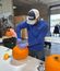 A man wearing a cap and blue shirt carves a pumpkin at a table during a fall activity in a workshop.
