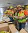 Workers in hard hats and safety vests stand in a break room line, holding plates of food at a lunch or appreciation event.