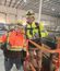 Two construction workers wearing high-visibility jackets, hard hats, safety glasses, and work gloves stand together on an orange lift inside a large industrial warehouse. Both are smiling and giving thumbs up to the camera.