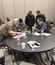 A group of men collaborates around a table, examining and adjusting a small wooden structure.