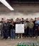 A large group of people stands together indoors holding a sign, with rows of canned goods arranged on the floor in front of them.