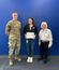 Three people pose in front of a blue wall, with the person in the center holding a framed certificate.