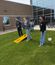 Three people stand on a grassy area outside a building, playing a bean bag toss game while others watch nearby.
