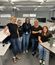 Five women in black shirts and jeans posing and smiling in a modern office cubicle area with computers, desks, and overhead industrial lighting.